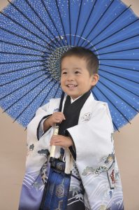 白地にさざ波と松竹柄の羽織に、青地の袴を着付けてもらいスタジオで撮影したこども。傘を持ち笑顔を浮かべている。着用しているのは三歳向けの七五三レンタル着物