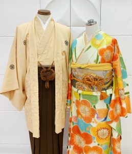 薄いベージュのメンズ着物に茶色の袴。クリームにオレンジ、黄緑、水色の梅が描写されたレディース衣装のナチュラルなコーデ