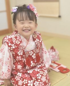 ﾋﾟﾝｸに桜尽くしの七五三用古典着物を着付けてもらい笑顔を浮かべる三歳の女の子