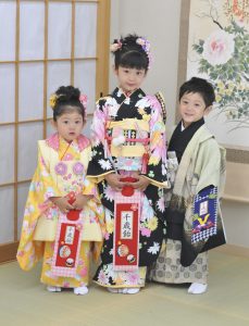 三歳五歳七歳の三人兄妹のお正月のような和服画像。姉は黒の四つ身、兄は黄緑のキッズサイズのメンズ紋服に袴。妹は黄色の被布をそれぞれ着付けてもらっている