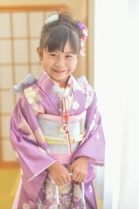 jillstuart風の藤の花っぽい綺 麗 な女の子七 五三用yotsumiを着 せてもらい笑顔を見せる少女。5歳～7歳ぐらいの子供が好む大人っぽい薄紫と水色の帯が顔によく映えている。窓口からの光が美しい