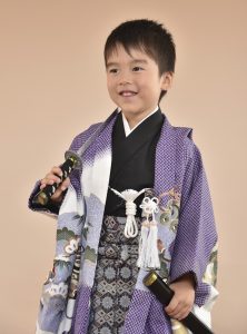 紫の正絹の様な疋田鹿の子のポリエステル素材に兜が描かれた５ 歳 着物を着付けられ撮影の小物を持った男の子七 五三思い出photo。シーズン中、引手数多な衣装は宅配が難しい
