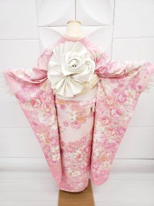 レディースの洋服で大人気なLIZLISAブランドのピンク色のバラ柄振袖。白無垢用の袋帯を複雑な薔薇の形に巻いている。帯結びがよく分かる袖を広げた後ろ姿の写真（桃パフェをイメージしてコーディネート）