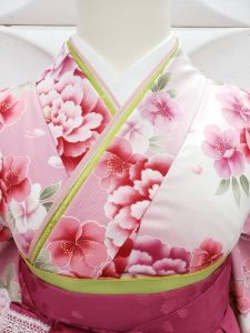 薄ピンクに牡丹の衣装に濃い桃色のぼかし、刺繍、レース入りの袴を着付けている。可愛らしいフルセットレンタルの一例。黄緑の重ね衿と半巾帯が確認可能