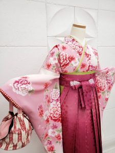 薄ピンクに牡丹の衣装に濃い桃色のぼかし、刺繍、レース入りの袴を着付けている。白に赤ピンクの矢絣巾着も可愛らしいフルセットレンタル。腰紐や襦袢、ブーツも届く