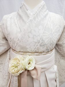 ベージュの菊柄小紋にアイボリーの無地袴のコーディネート。花とレースの飾りが結び目に付けられている。卒業式の袴でワントーンコーデをした一例。胸元のアップで帯や重ね衿も見える
