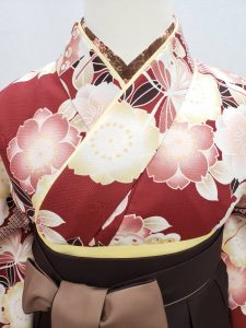 赤茶に八重桜と蝶の花の丸が描かれた絞り風二尺袖。袴は焦げ茶で内側と紐がベージュ。同じトーンでまとまったレトロモダンで落ち着いた卒業式のフルセット。茶色の半襟や黄色の重ね衿がよく見える