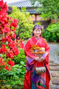 花と緑が溢れる日本庭園で赤色のママ振袖を着付けられた姿で前撮りをしている成人を迎える女の子。髪型は水引きと金箔を使った和装ヘアアレンジ