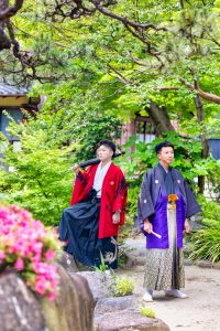 緑豊かな日本庭園で赤の羽織りに黒の袴の成人者と紫に変わる黒地の紋付きに白系の袴姿の男の子二人組みが背中合わせに立った前撮り写真