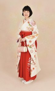 白地の可愛らしい振袖に朱赤の刺繍入り袴、ピンクの帯を着付けた大学卒業の女性。和装用の丸く桜色のバックを持っている
