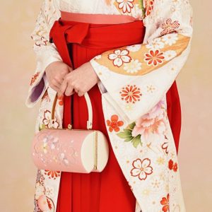 白地の可愛らしい振袖に朱赤の刺繍入り袴、ピンクの帯を着付けた大学卒業の女性。和装用の丸く桜色のバックを持っている